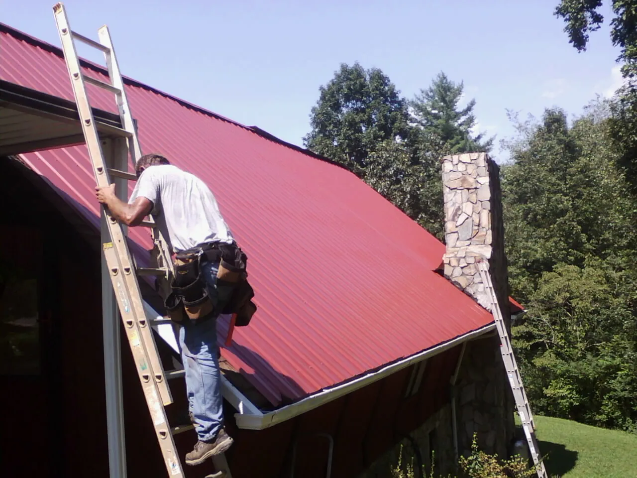 Roofer on a ladder beside a red metal roof and stone chimney — Soffit & Fascia Repair by Roofing Brothers in Hartleton